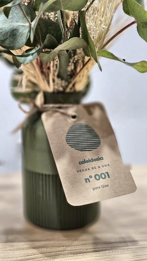 Florero verde con flores secas y la etiqueta de Caleidosia: hecha de a una, nº 001